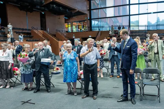 Uczestnicy wydarzenia z bukietami kwiatów, świętujący w sali z krzesłami.