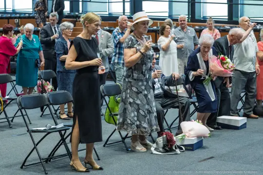 Grupa osób w sali, niektórzy trzymają kwiaty, inni tańczą lub klaskają.