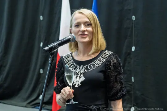 Kobieta w eleganckiej sukience trzyma kieliszek, przemawia do zgromadzonej publiczności.