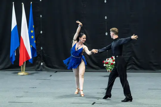 Para taneczna w strojach balowych tańczy na scenie, trzymając się za ręce.