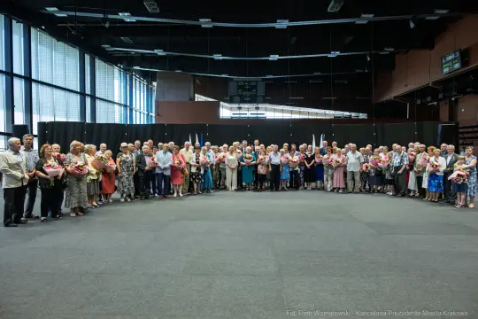 Grupa ludzi stojąca w sali, trzymająca bukiety kwiatów.