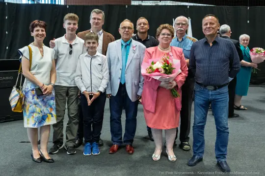Grupa osób pozująca do zdjęcia, w tym mężczyzna w jasnym garniturze i kobieta z bukietem kwiatów.