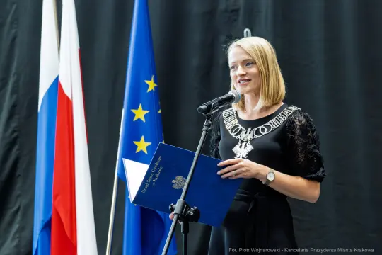 Kobieta przemawia do mikrofonu, trzymając dokument, w tle flagi Rosji i Unii Europejskiej.
