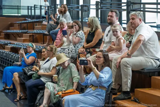 Grupa ludzi siedzących na widowni, niektórzy trzymają telefony, inni rozmawiają.