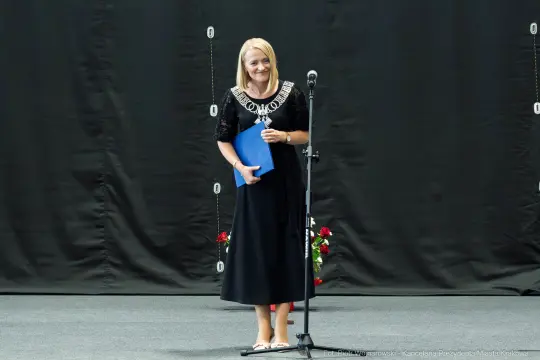 Kobieta w czarnej sukni z uśmiechem trzyma niebieski dokument przy mikrofonie.