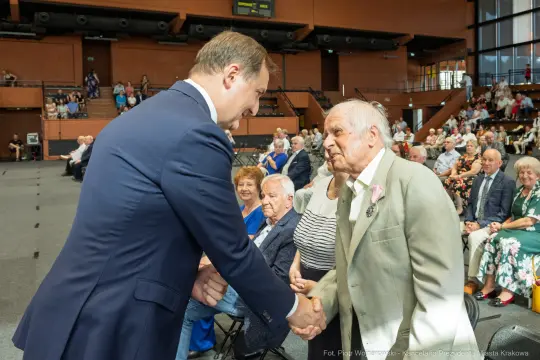 Spotkanie młodszego mężczyzny z starszym, uściski dłoni, w tle publiczność.