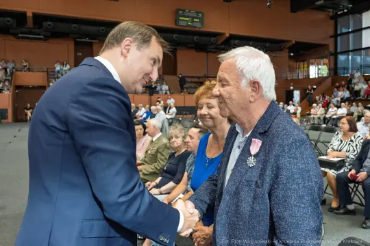 Mężczyzna w garniturze wita się z starszym mężczyzną, obok stoi kobieta. W tle widownia.