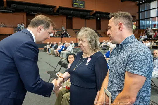 Uroczystość wręczenia odznaczeń, mężczyzna podaje rękę kobiecie z medalem.