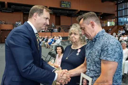 Mężczyzna w garniturze ściska dłoń innego mężczyzny, w tle siedzący tłum ludzi.
