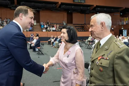 Spotkanie trzech osób, w tym mężczyzny w garniturze i dwóch starszych ludzi, w sali z widownią.