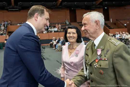 Uścisk dłoni między mężczyzną a starszym mężczyzną w mundurze, obok kobieta w różowej sukience.