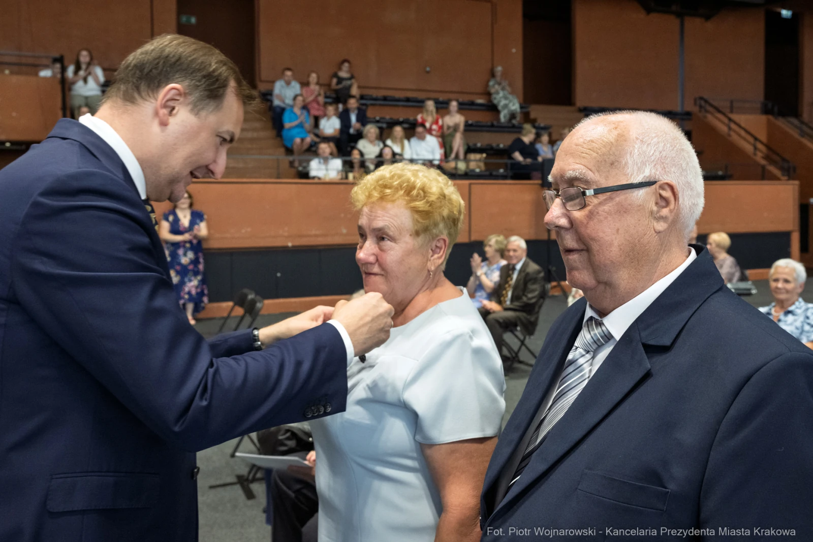Wręczenie odznaczenia kobiecie przez mężczyznę w eleganckim otoczeniu, obok stoi starszy mężczyzna.  Autor: Piotr Wojnarowski