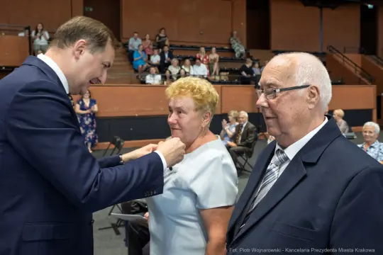 Wręczenie odznaczenia kobiecie przez mężczyznę w eleganckim otoczeniu, obok stoi starszy mężczyzna.