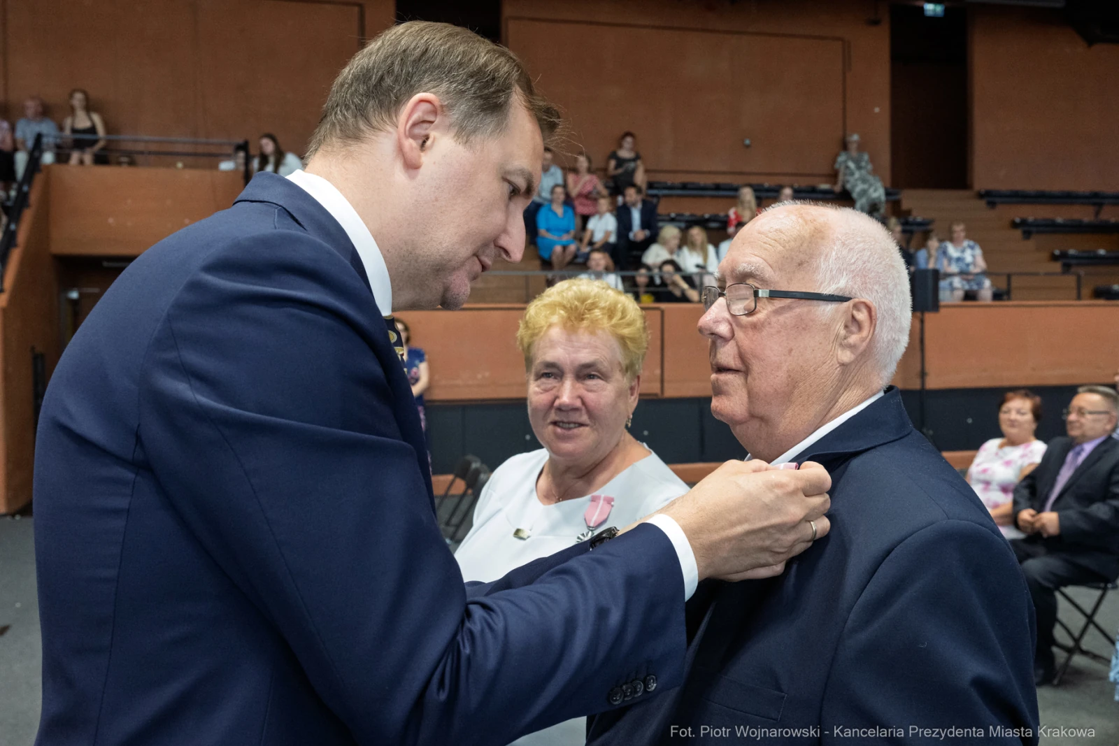 Uroczystość wręczenia odznaczenia mężczyźnie w garniturze przez innego mężczyznę. W tle kobieta i publiczność.  Autor: Piotr Wojnarowski