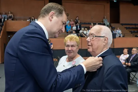 Uroczystość wręczenia odznaczenia mężczyźnie w garniturze przez innego mężczyznę. W tle kobieta i publiczność.