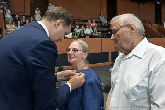 Wręczenie odznaczenia kobiecie przez mężczyznę w obecności starszego mężczyzny.