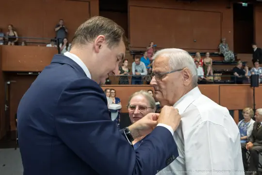Wręczenie odznaczenia mężczyźnie przez innego mężczyznę w sali pełnej osób.