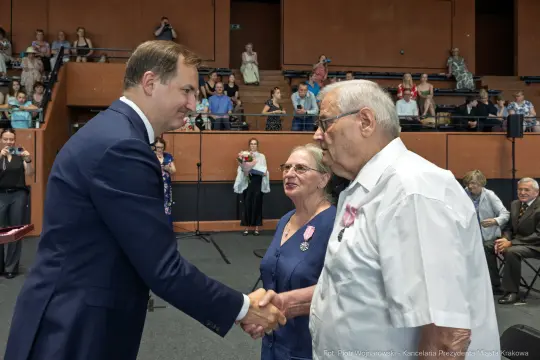Uroczystość wręczenia odznaczeń, mężczyzna w garniturze podaje rękę starszemu mężczyźnie.