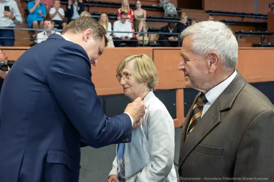 Wręczenie odznaczenia przez mężczyznę kobiecie w towarzystwie starszego mężczyzny.