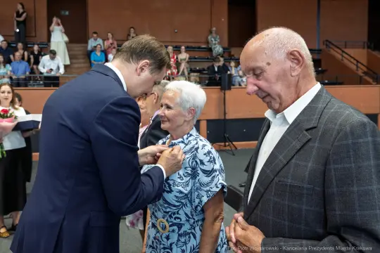 Wręczenie odznaczenia starszej kobiecie przez mężczyznę w garniturze, obok stoi starszy mężczyzna.