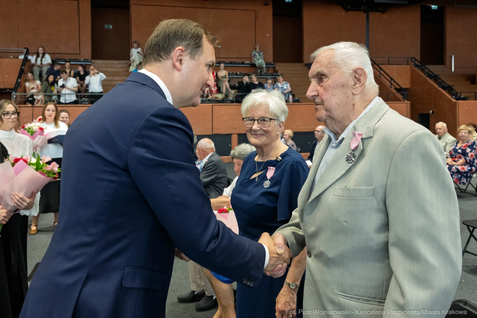 Mężczyzna w garniturze ściska dłoń starszemu mężczyźnie, w tle kobieta z bukietem kwiatów.  Autor: Piotr Wojnarowski