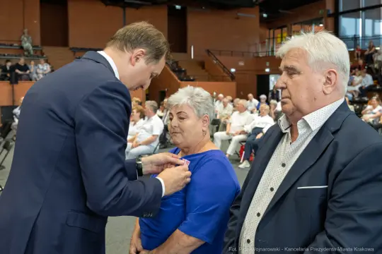 Wręczenie odznaczenia przez mężczyznę kobiecie w obecności innego mężczyzny.