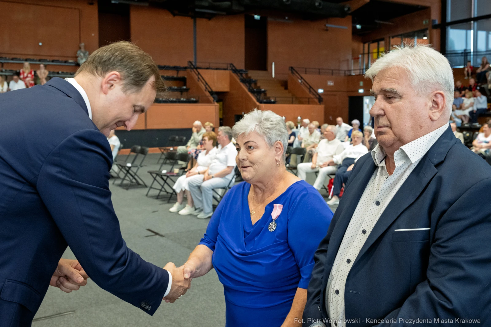 Uroczystość wręczenia odznaczeń, mężczyzna podaje rękę kobiecie w niebieskiej sukience.  Autor: Piotr Wojnarowski