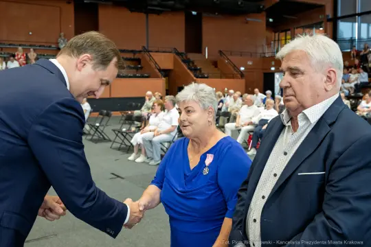 Uroczystość wręczenia odznaczeń, mężczyzna podaje rękę kobiecie w niebieskiej sukience.