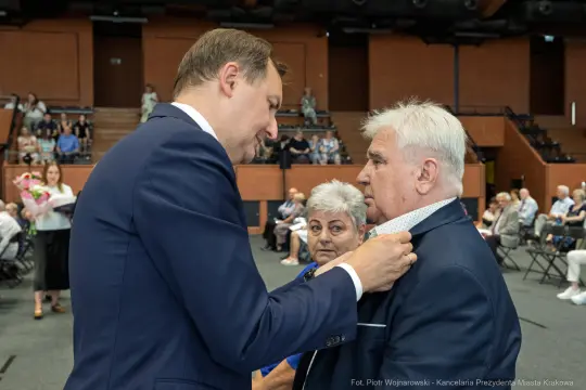 Wręczenie odznaczenia mężczyźnie przez innego mężczyznę w obecności kobiety.