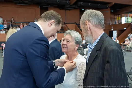 Wręczenie odznaczenia kobiecie przez mężczyznę w formalnym stroju, obok stoi starszy mężczyzna.