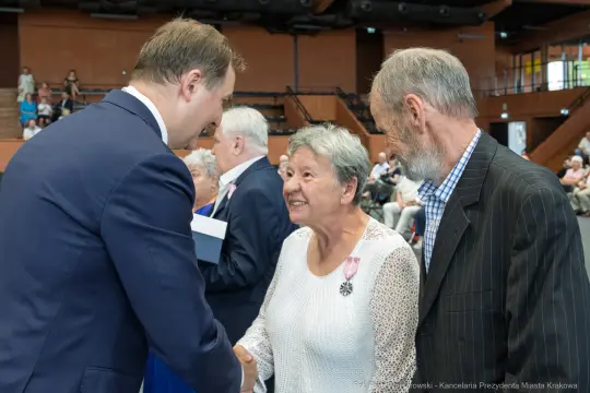 Spotkanie trzech osób, w tym uśmiechniętej kobiety w jasnej sukience, podczas wręczenia odznaczeń.