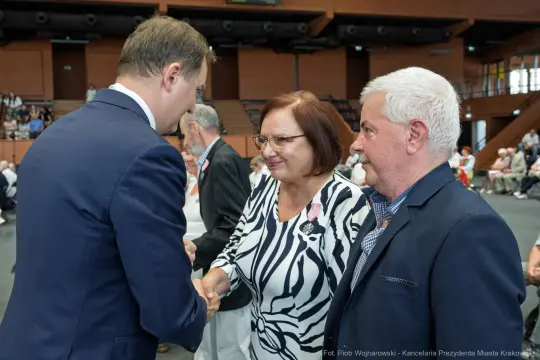 Spotkanie trzech osób, jedna z nich podaje rękę drugiej. W tle widoczna publiczność.