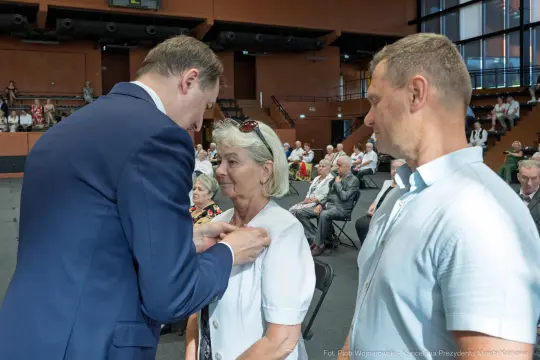 Wręczenie odznaczenia kobiecie przez mężczyznę w obecności innych uczestników wydarzenia.