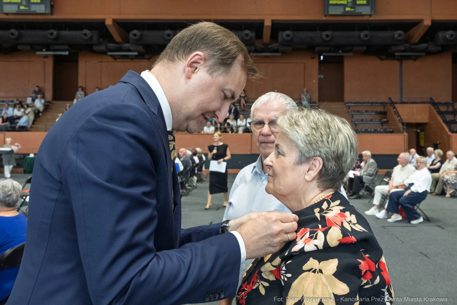 Mężczyzna przypina odznaczenie kobiecie w eleganckim stroju, w tle zgromadzenie osób.  Autor: Piotr Wojnarowski