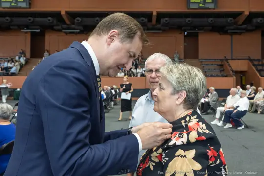 Mężczyzna przypina odznaczenie kobiecie w eleganckim stroju, w tle zgromadzenie osób.