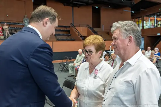 Uroczystość wręczenia odznaczeń, mężczyzna w garniturze wita się z parą starszych osób.