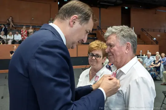 Wręczenie odznaczeń przez mężczyznę starszej parze w sali pełnej widzów.