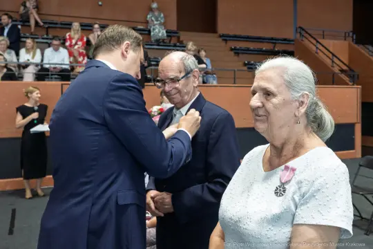 Wręczenie odznaczenia mężczyźnie przez uśmiechniętego mężczyznę, obok stoi kobieta.