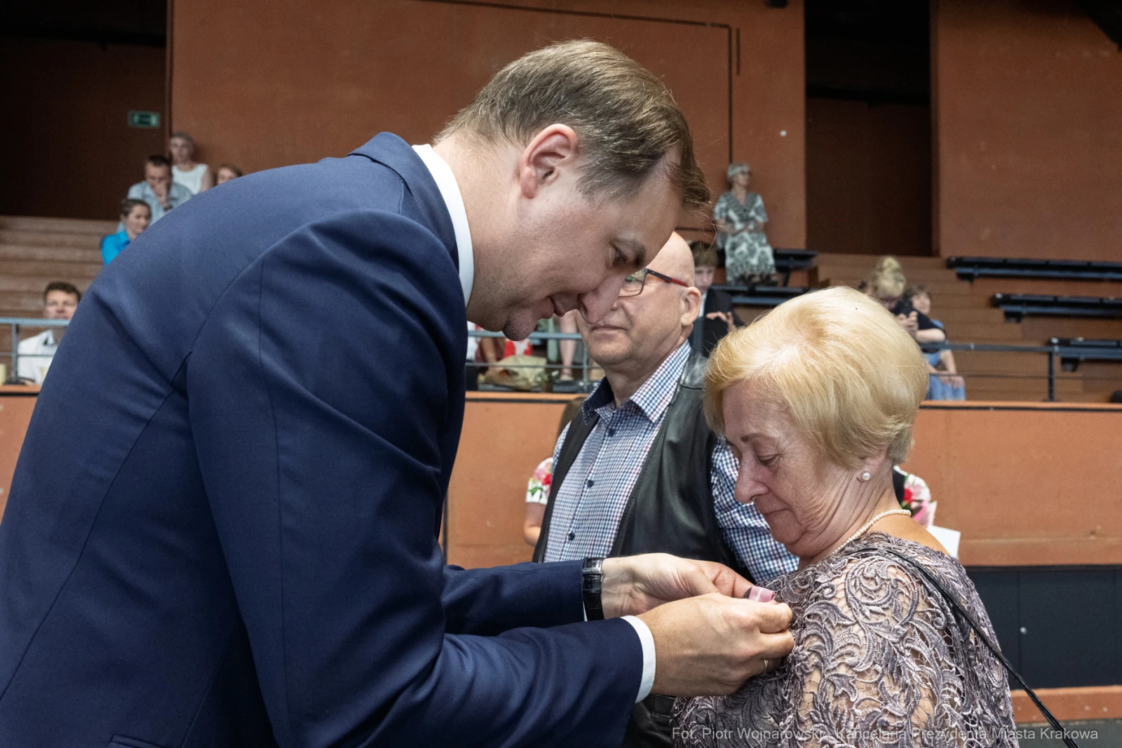 Mężczyzna przypina odznaczenie do ubrania kobiety w towarzystwie innych osób.  Autor: Piotr Wojnarowski