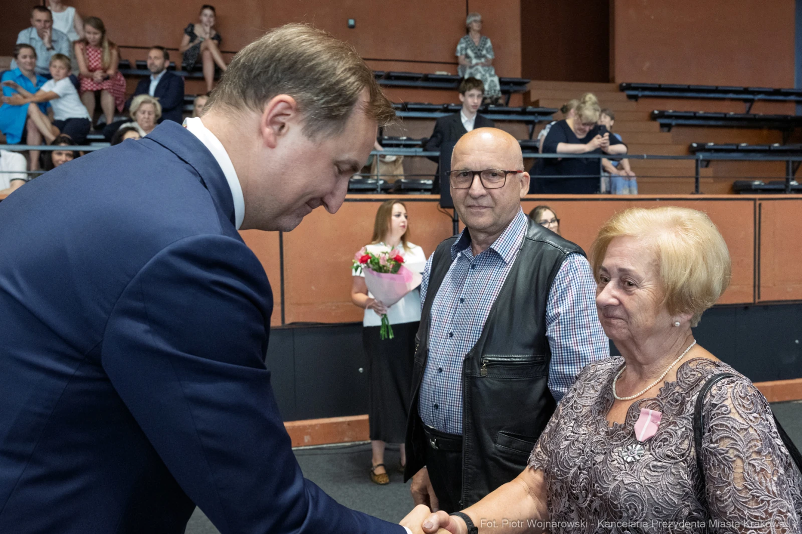 Uroczystość wręczenia nagród, mężczyzna w garniturze podaje rękę starszemu mężczyźnie. Kobieta z bukietem kwiatów  Autor: Piotr Wojnarowski