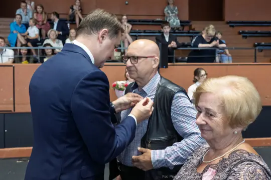 Wręczenie odznaczenia mężczyźnie przez elegancko ubranego mężczyznę, w tle widownia.