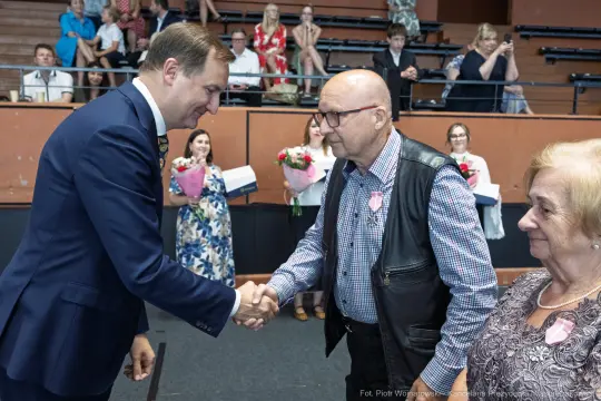 Uścisk dłoni między mężczyzną w garniturze a starszym mężczyzną w kamizelce, w tle goście z kwiatami.