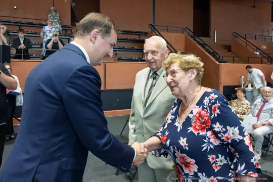 Mężczyzna w garniturze podaje rękę starszej kobiecie, obok stoi starszy mężczyzna.
