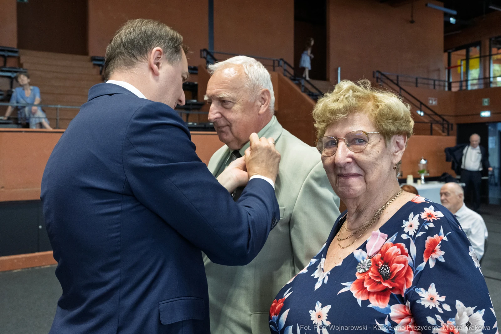 Mężczyzna przypina odznaczenie starszemu mężczyźnie, obok stoi uśmiechająca się kobieta.  Autor: Piotr Wojnarowski