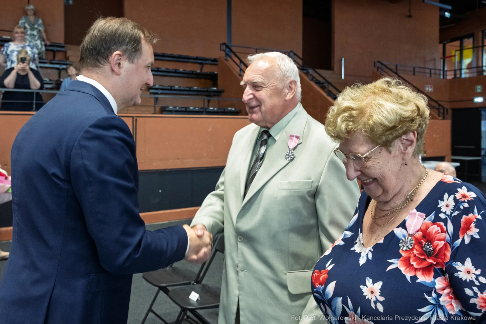 Uroczystość wręczenia odznaczeń, mężczyzna w garniturze wita starszych państwa.  Autor: Piotr Wojnarowski