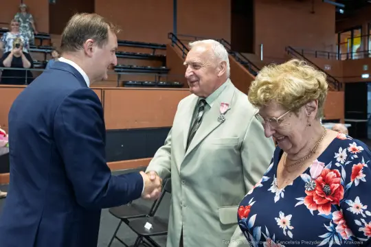Uroczystość wręczenia odznaczeń, mężczyzna w garniturze wita starszych państwa.