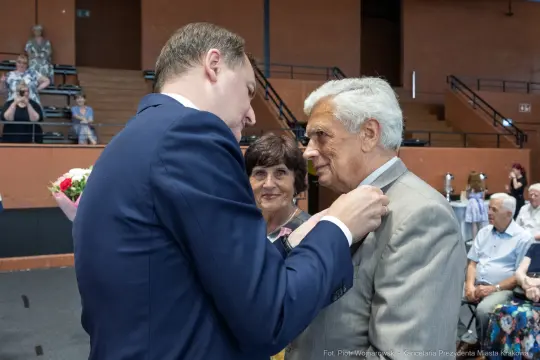 Mężczyzna przypina odznaczenie starszemu mężczyźnie, obok stoi kobieta.