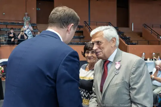 Mężczyzna w garniturze rozmawia z uśmiechniętym starszym panem w odznaczeniem. W tle widoczni goście.