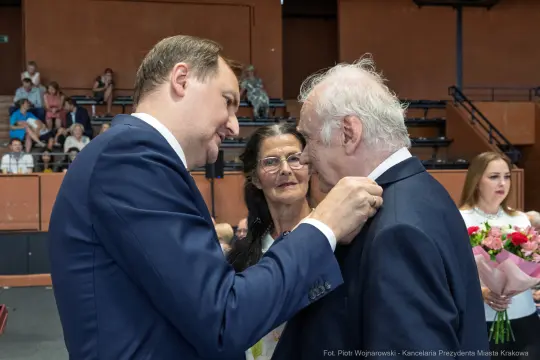 Mężczyzna przypina odznaczenie starszemu mężczyźnie, w tle kobieta z bukietem kwiatów.