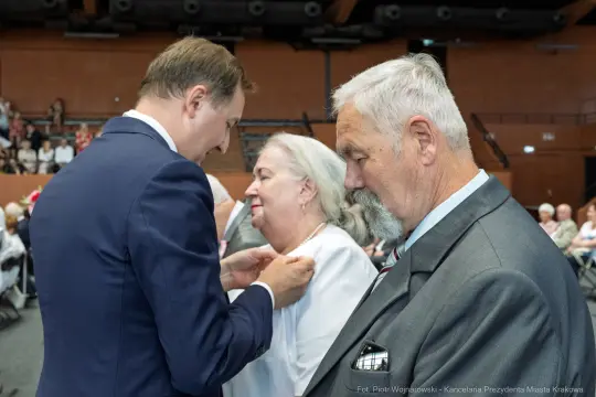 Wręczenie odznaczenia przez mężczyznę kobiecie w obecności starszego mężczyzny.